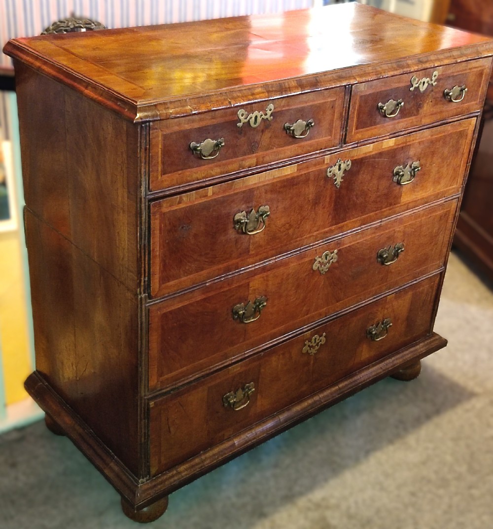 An early eighteenth-century walnut chest of drawers featuring decorative brass handles, crossbanded drawer fronts, and turned bun feet, constructed in two separate upper and lower sections. View VII