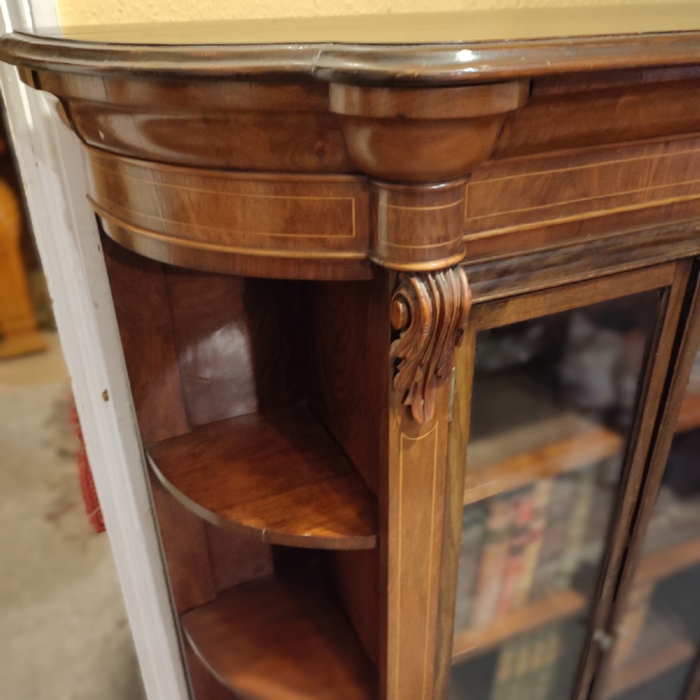 An antique Victorian walnut glazed bookcase featuring double glass doors flanked by open curved side shelves and carved corbels. View II