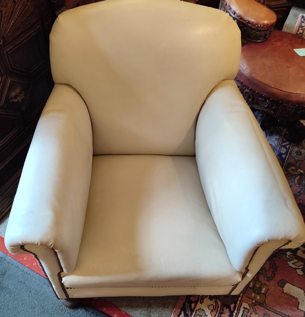 A cream leather Edwardian club chair featuring brass studding along the front edges and resting on short turned wooden feet. View II