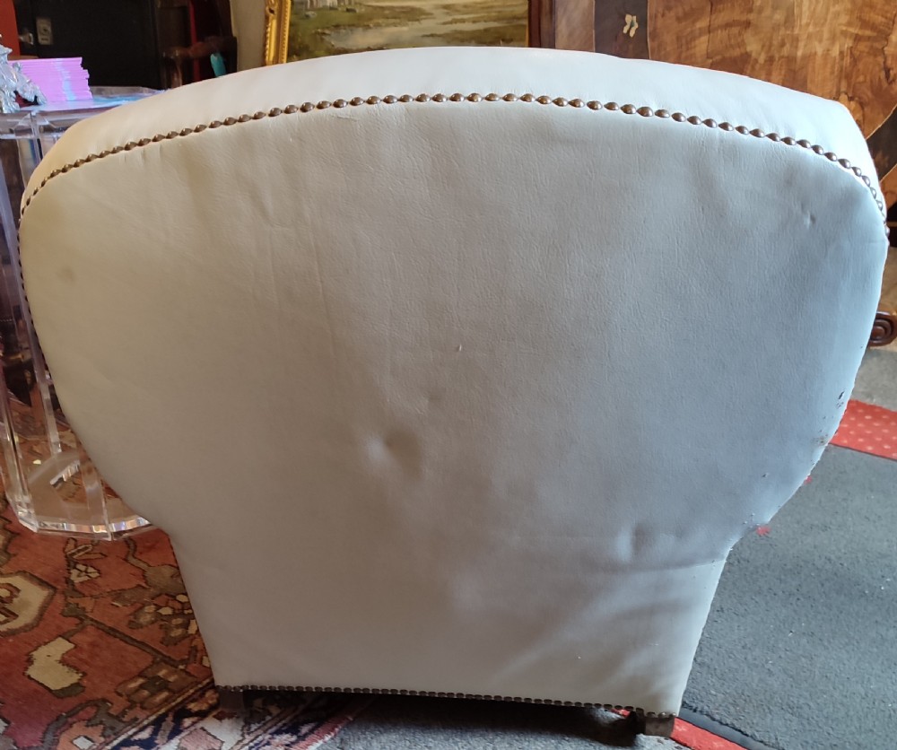 A cream leather Edwardian club chair featuring brass studding along the front edges and resting on short turned wooden feet. View V