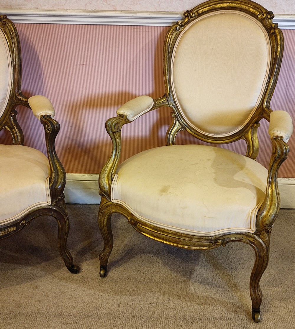 A pair of 19th-century giltwood armchairs featuring carved gold frames, oval backrests, padded arms, cabriole legs, and light cream upholstery. View II