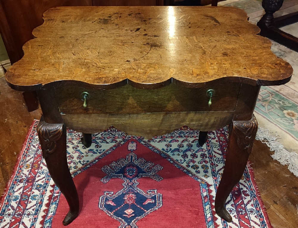 An 18th-century oak lowboy featuring a scalloped edge top, a single drawer with brass drop handles, a shaped apron, and carved cabriole legs.