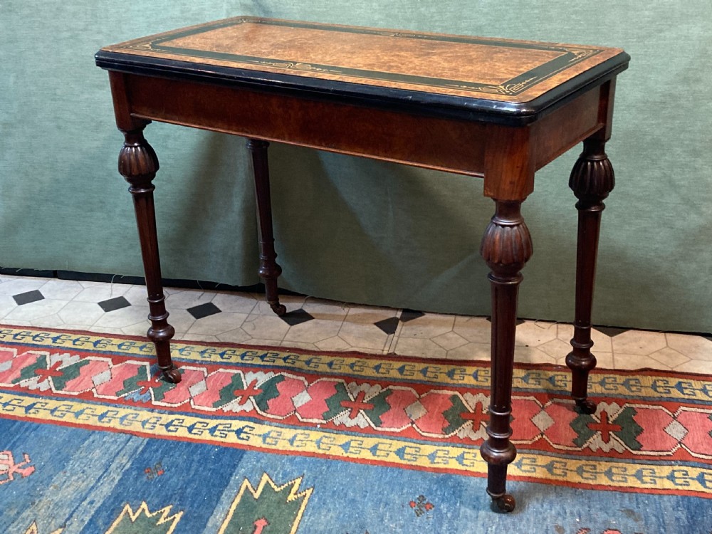 A Victorian burr walnut fold-over card table featuring an ebonised top border with gilt detailing and four turned, fluted legs set on brass castors.