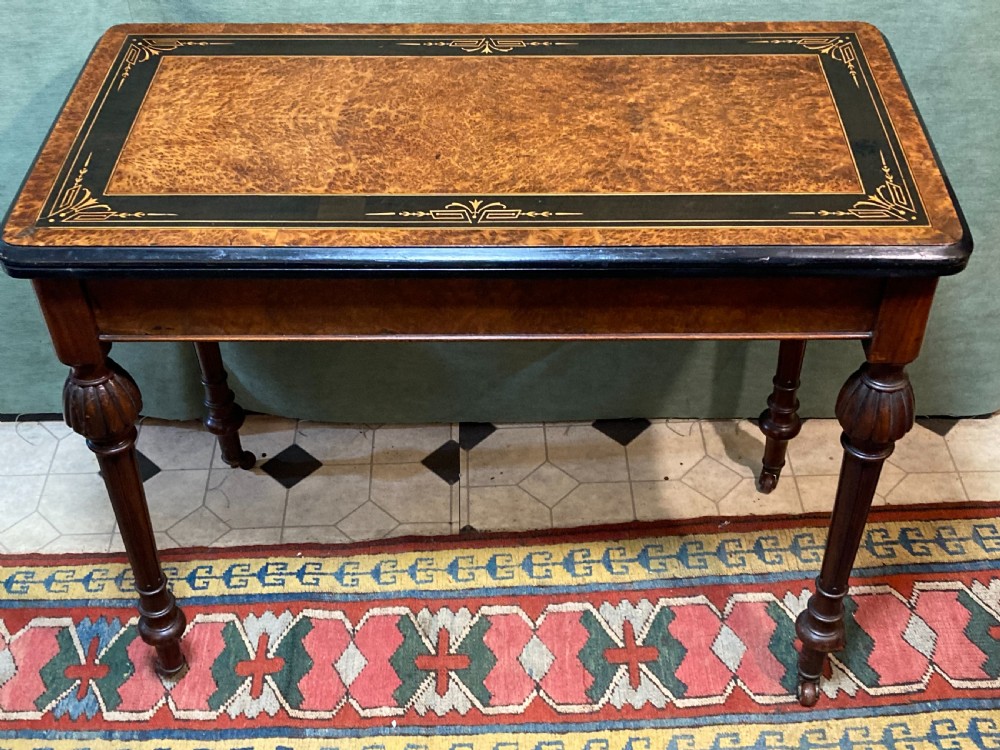 A Victorian burr walnut fold-over card table featuring an ebonised top border with gilt detailing and four turned, fluted legs set on brass castors. View II