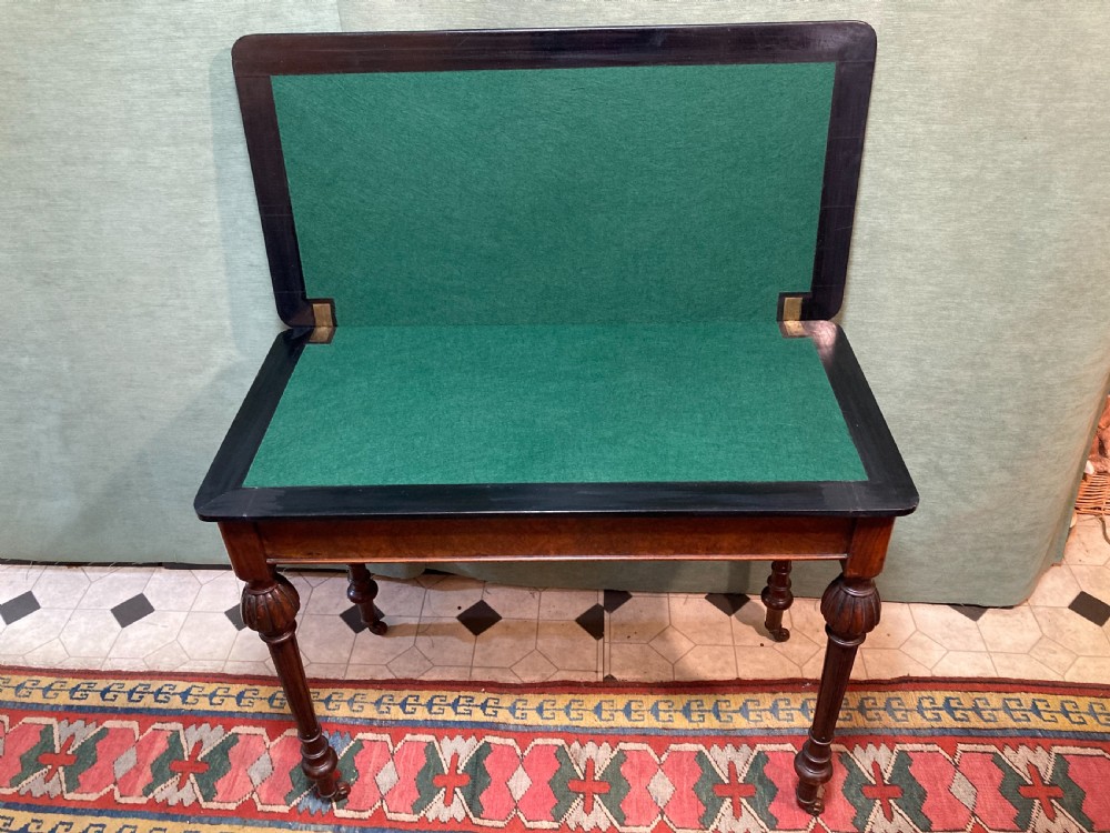 A Victorian burr walnut fold-over card table featuring an ebonised top border with gilt detailing and four turned, fluted legs set on brass castors. View III
