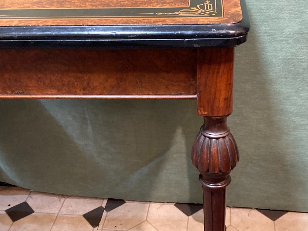 A Victorian burr walnut fold-over card table featuring an ebonised top border with gilt detailing and four turned, fluted legs set on brass castors. View IV