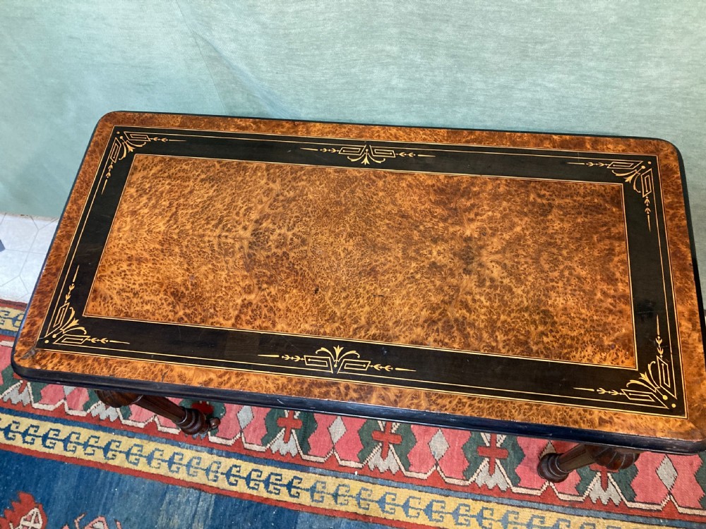 A Victorian burr walnut fold-over card table featuring an ebonised top border with gilt detailing and four turned, fluted legs set on brass castors. View V