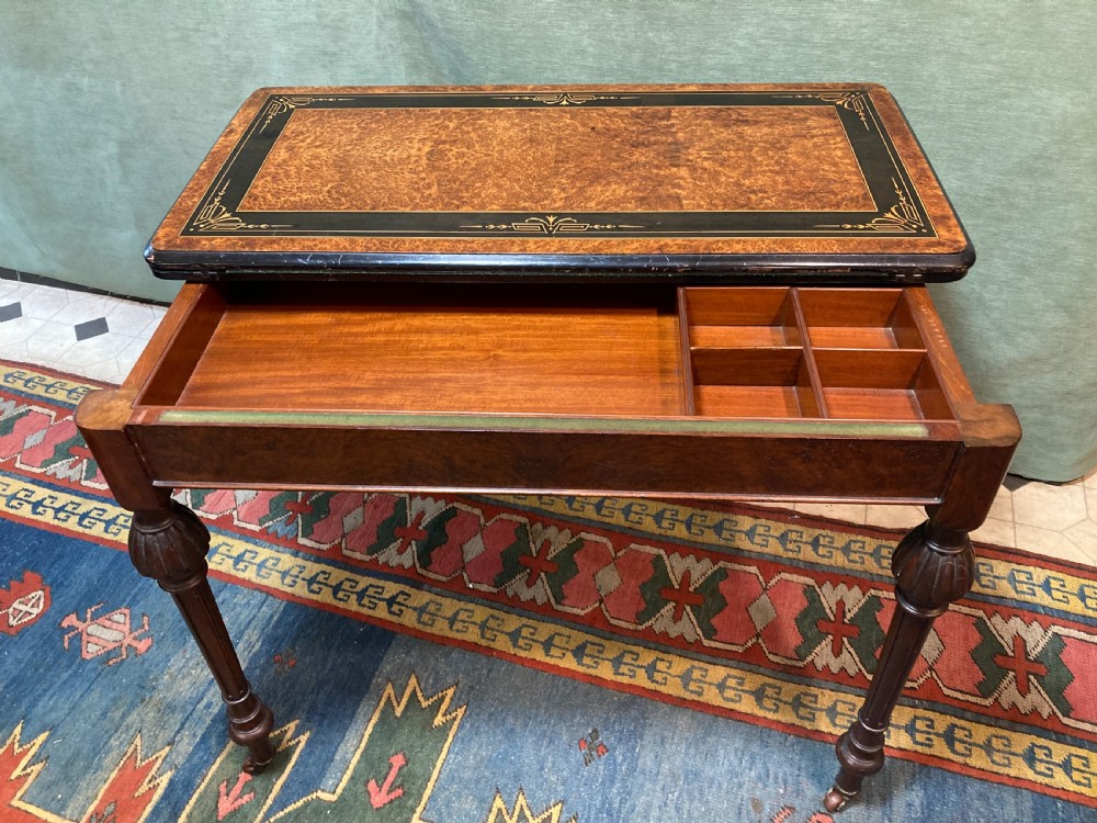 A Victorian burr walnut fold-over card table featuring an ebonised top border with gilt detailing and four turned, fluted legs set on brass castors. View VI