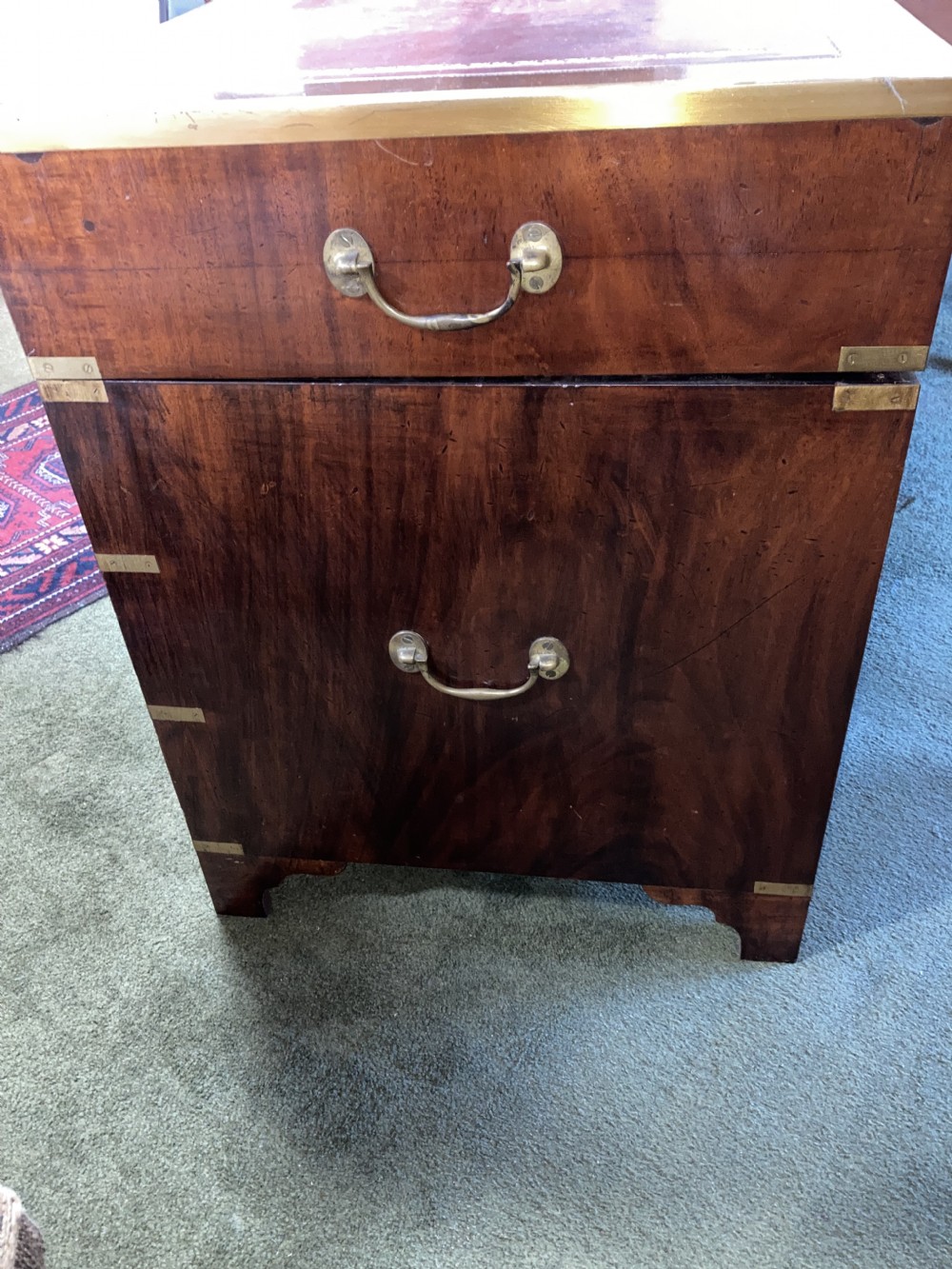 An Edwardian mahogany campaign pedestal desk featuring flush-fitting brass handles, protective brass corner mounts, and a red leather inset writing surface. View VI