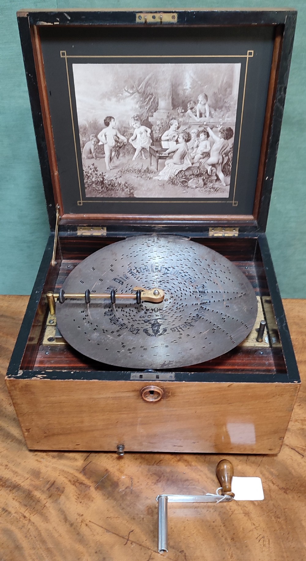 An open 19th-century walnut Polyphon music box featuring a metal disc, central pressure arm, winding crank, and a monochrome print of cherubs on the underside of the lid.