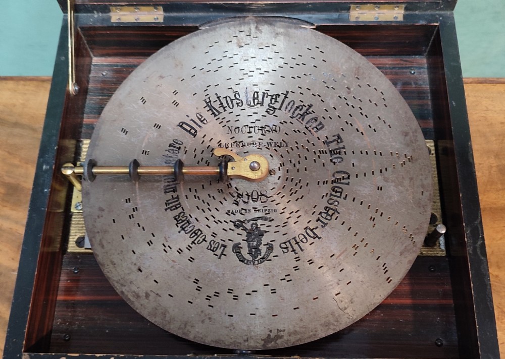 An open 19th-century walnut Polyphon music box featuring a metal disc, central pressure arm, winding crank, and a monochrome print of cherubs on the underside of the lid. View III