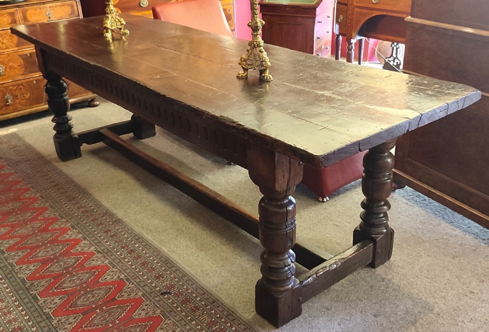 A long dark oak refectory dining table featuring a carved aproned border, stout turned legs, and a low rectangular stretcher base. View II