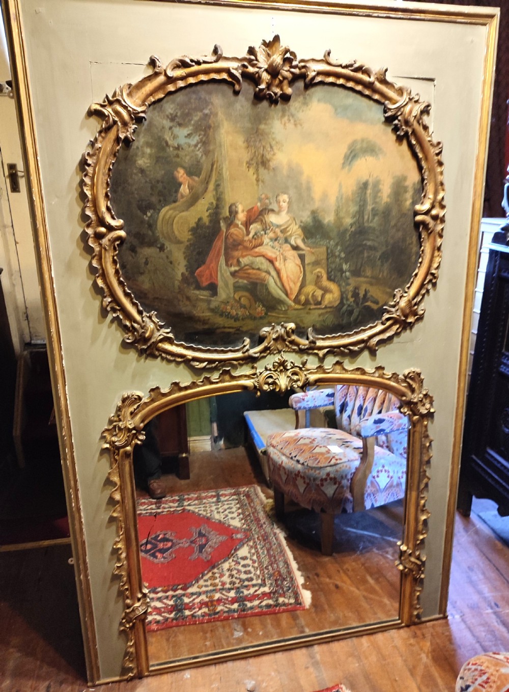 A large nineteenth-century French trumeau mirror featuring a painted canvas of a romantic pastoral scene above a mirror glass, set within carved giltwood frames on a grey painted panel.