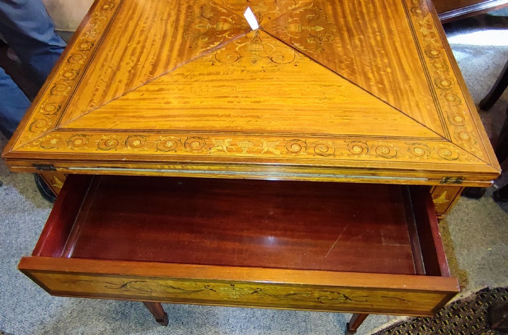 An Edwardian satinwood envelope card table featuring detailed marquetry inlays, four triangular top flaps, a single frieze drawer, and square tapered legs with a lower undertier. View VII