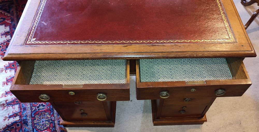 A Victorian wooden pedestal desk featuring a red leather inset top, brass ring handles, and multiple drawers supported on plinth bases. View V