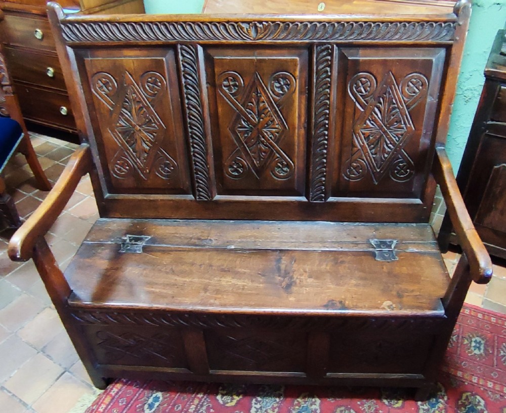 An early 18th-century dark oak box settle featuring three carved back panels, raised armrests, and a hinged lid seat for storage.