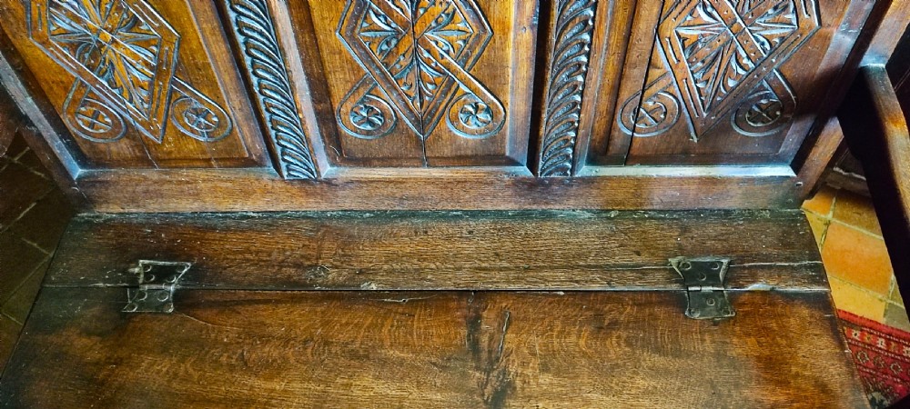 An early 18th-century dark oak box settle featuring three carved back panels, raised armrests, and a hinged lid seat for storage. View III