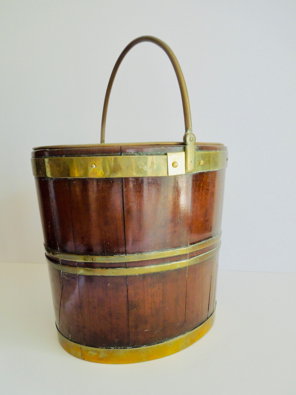 An oval mahogany wine bucket constructed from timber staves, bound by three polished brass hoops, and fitted with an arched brass swing handle.