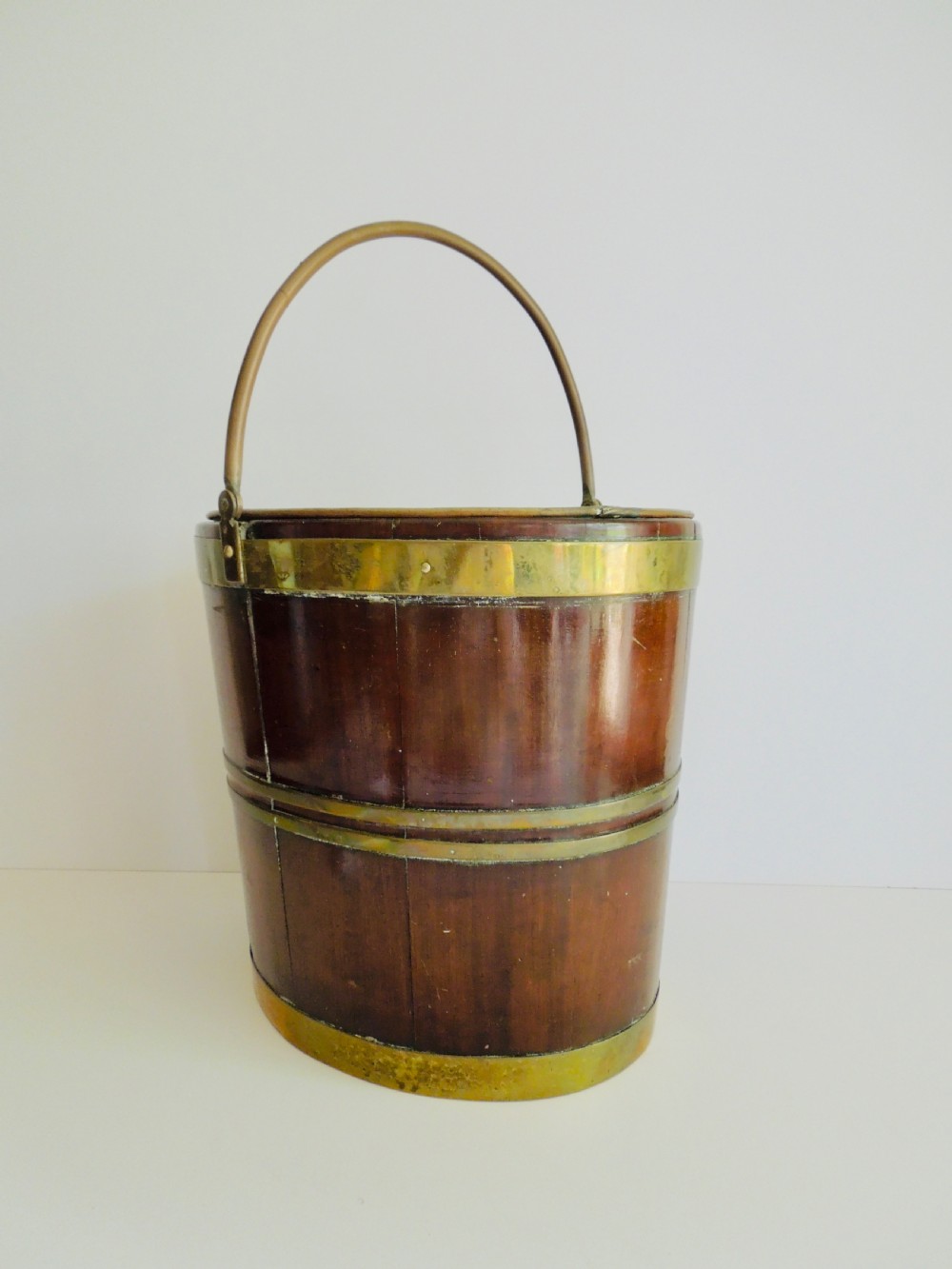 An oval mahogany wine bucket constructed from timber staves, bound by three polished brass hoops, and fitted with an arched brass swing handle. View II