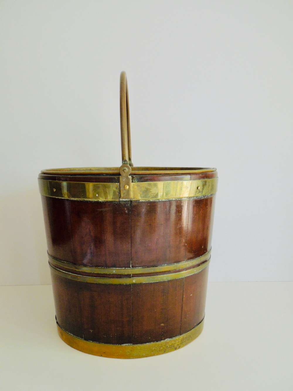 An oval mahogany wine bucket constructed from timber staves, bound by three polished brass hoops, and fitted with an arched brass swing handle. View III