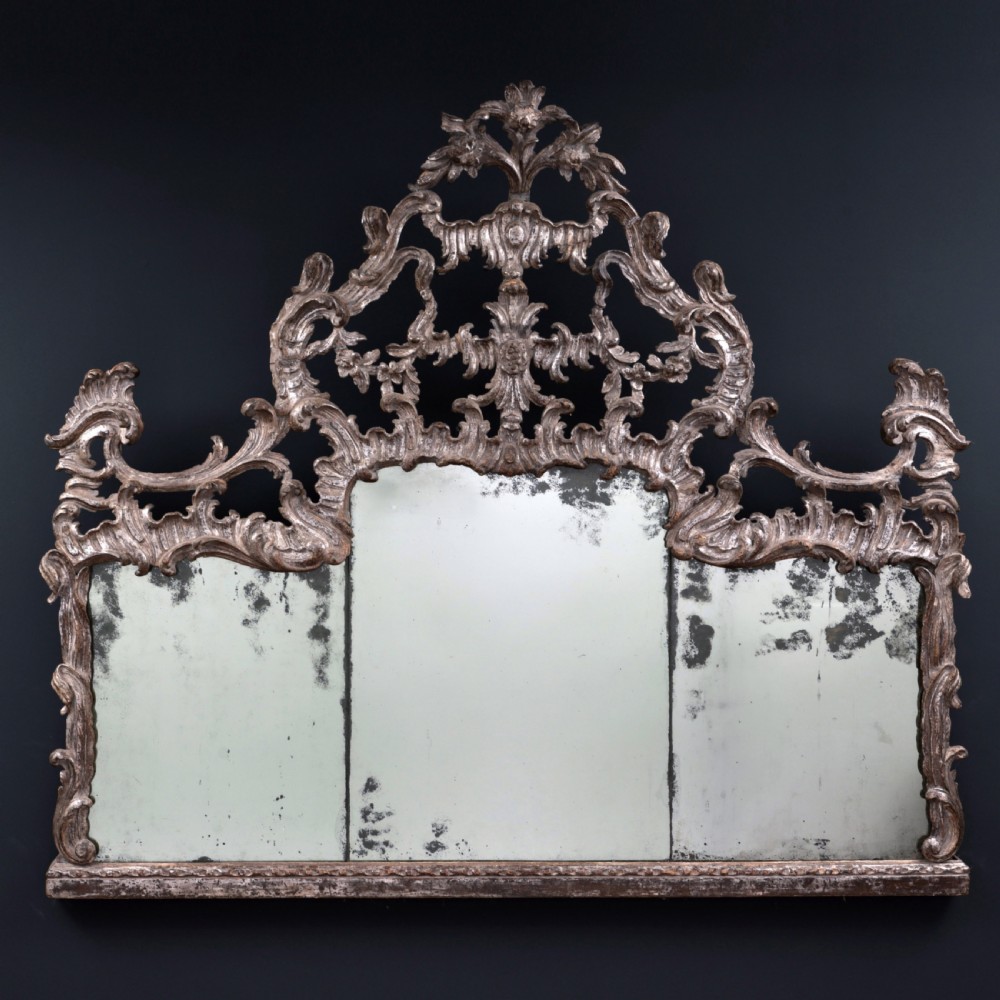 An 18th-century silvered Rococo overmantel mirror featuring an elaborately carved crest with C-scrolls and a tripartite mirror plate with visible foxing.