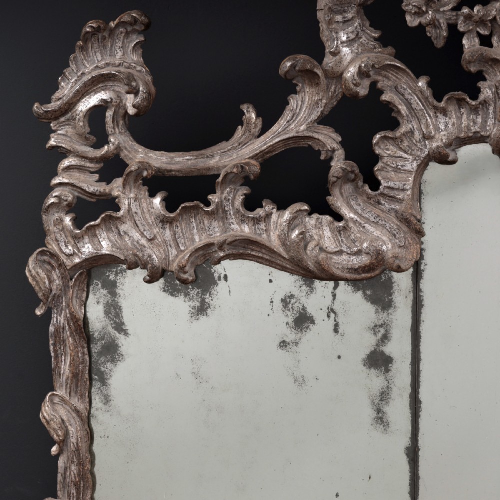 An 18th-century silvered Rococo overmantel mirror featuring an elaborately carved crest with C-scrolls and a tripartite mirror plate with visible foxing. View II