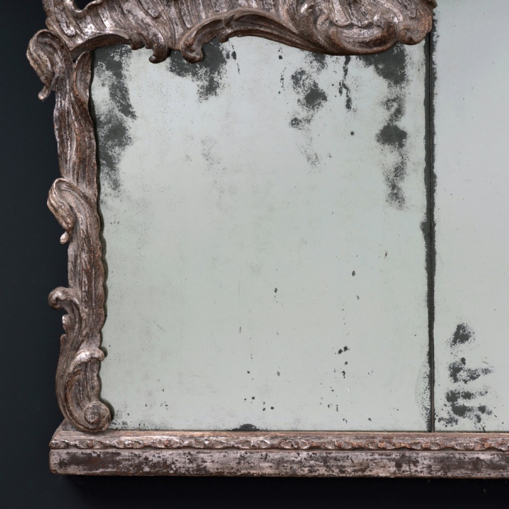 An 18th-century silvered Rococo overmantel mirror featuring an elaborately carved crest with C-scrolls and a tripartite mirror plate with visible foxing. View V