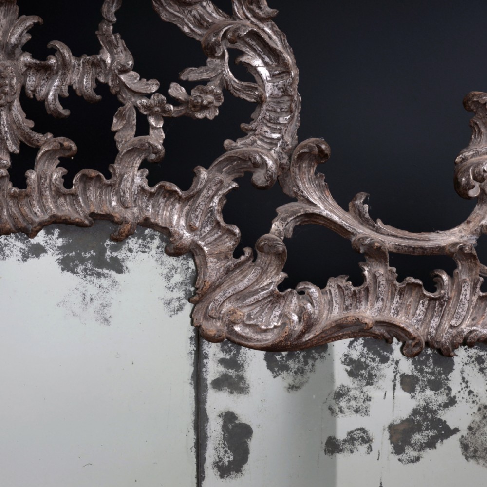 An 18th-century silvered Rococo overmantel mirror featuring an elaborately carved crest with C-scrolls and a tripartite mirror plate with visible foxing. View VI
