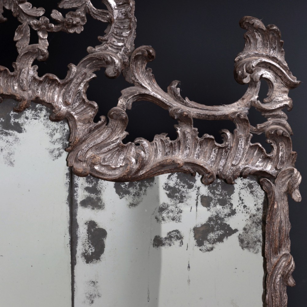An 18th-century silvered Rococo overmantel mirror featuring an elaborately carved crest with C-scrolls and a tripartite mirror plate with visible foxing. View VII