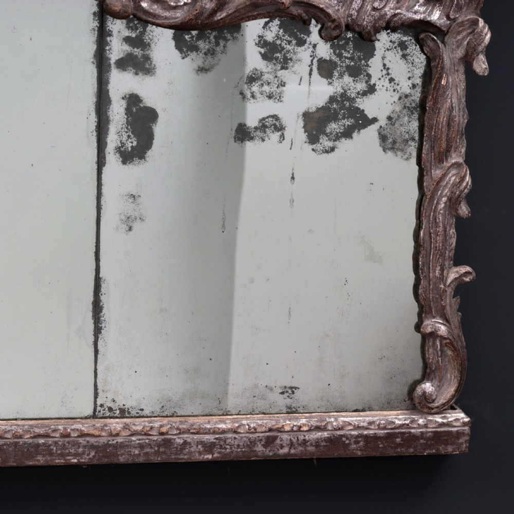An 18th-century silvered Rococo overmantel mirror featuring an elaborately carved crest with C-scrolls and a tripartite mirror plate with visible foxing. View VIII