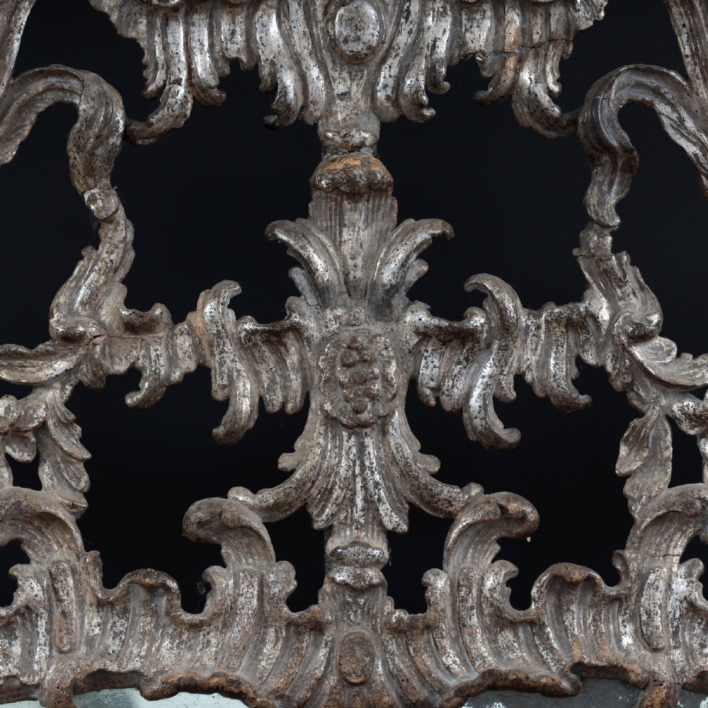 An 18th-century silvered Rococo overmantel mirror featuring an elaborately carved crest with C-scrolls and a tripartite mirror plate with visible foxing. View IX