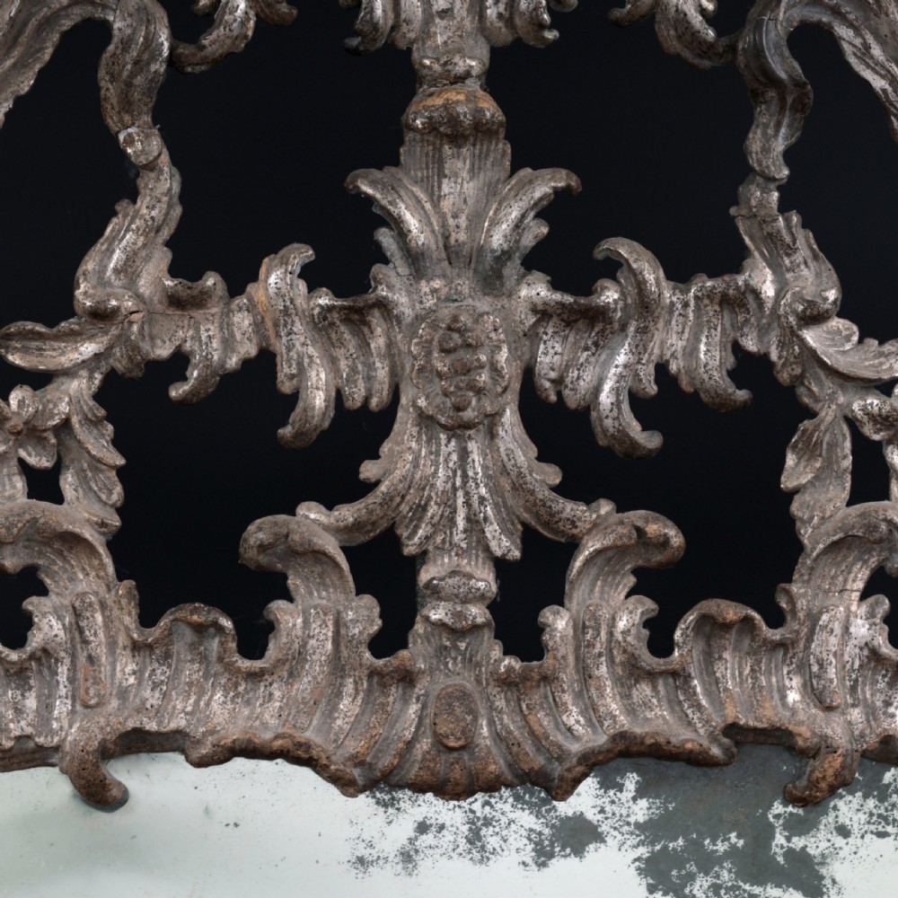 An 18th-century silvered Rococo overmantel mirror featuring an elaborately carved crest with C-scrolls and a tripartite mirror plate with visible foxing. View X