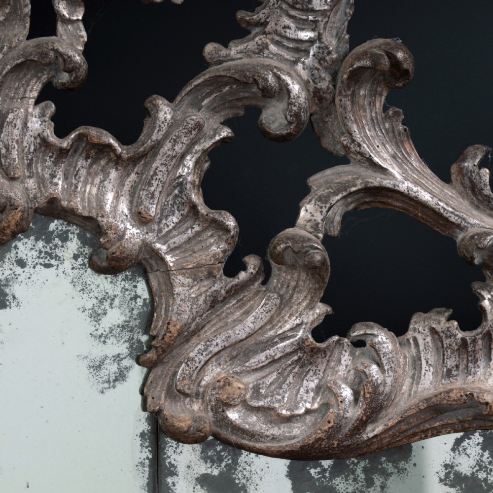 An 18th-century silvered Rococo overmantel mirror featuring an elaborately carved crest with C-scrolls and a tripartite mirror plate with visible foxing. View XII