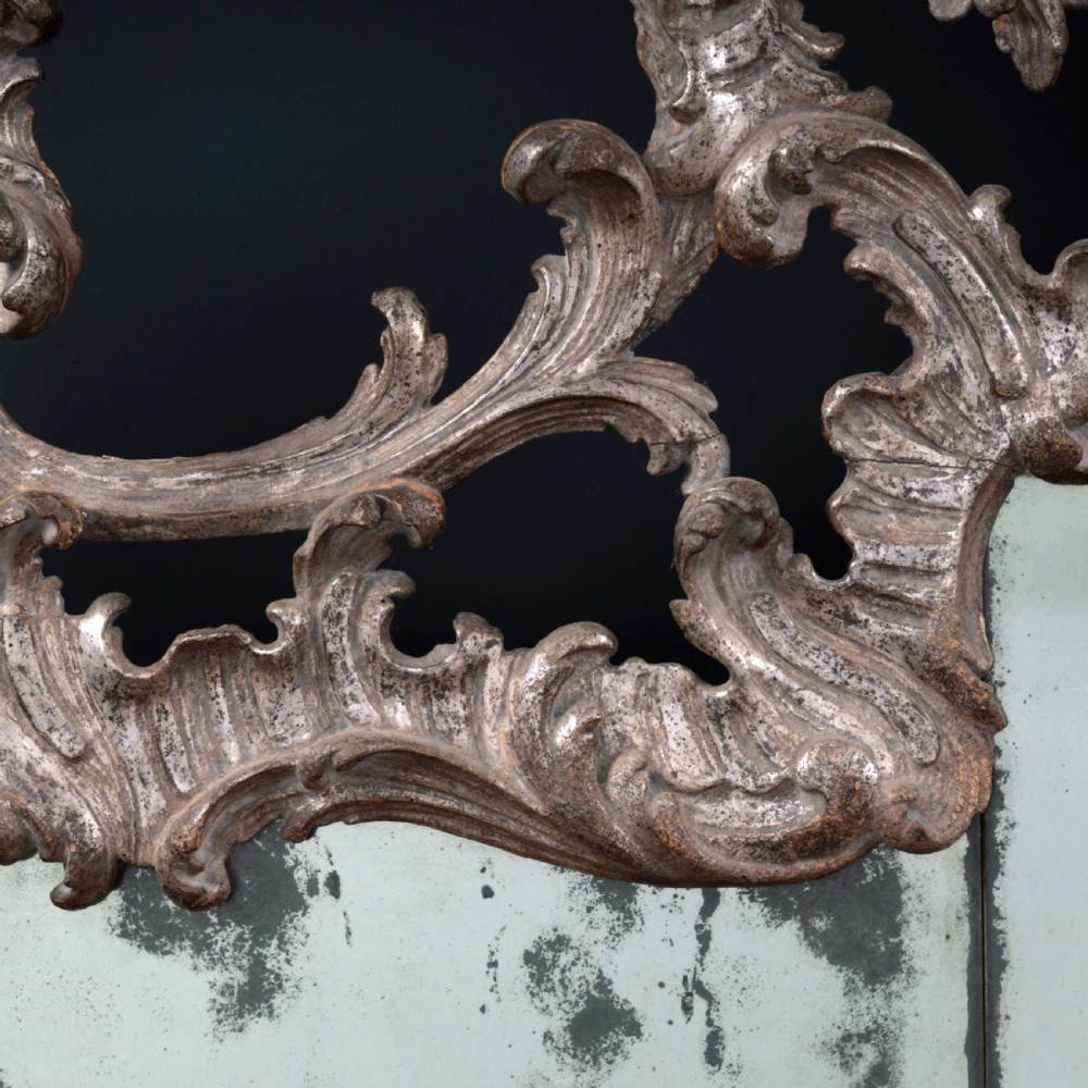 An 18th-century silvered Rococo overmantel mirror featuring an elaborately carved crest with C-scrolls and a tripartite mirror plate with visible foxing. View XIII