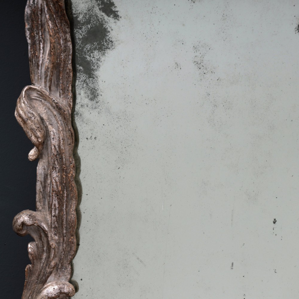An 18th-century silvered Rococo overmantel mirror featuring an elaborately carved crest with C-scrolls and a tripartite mirror plate with visible foxing. View XIV