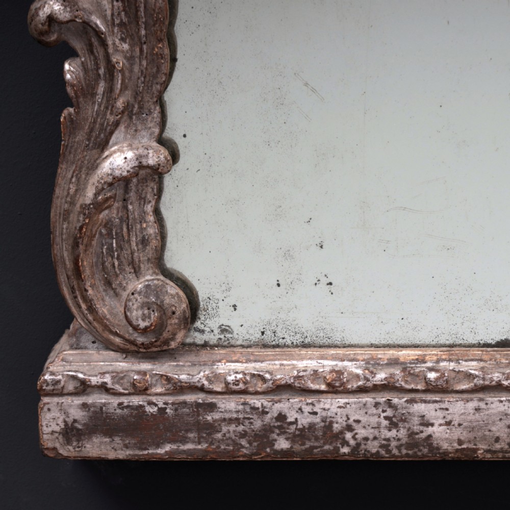 An 18th-century silvered Rococo overmantel mirror featuring an elaborately carved crest with C-scrolls and a tripartite mirror plate with visible foxing. View XV