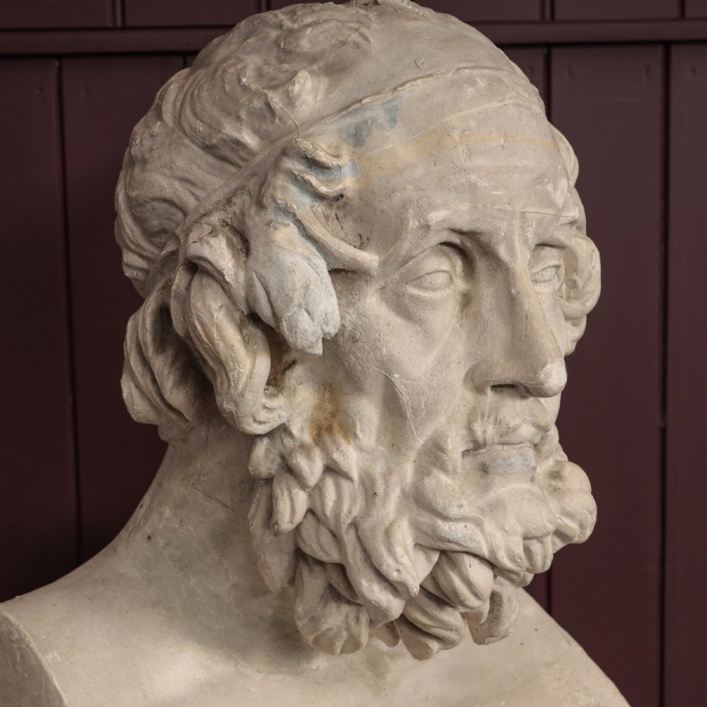 An off-white plaster bust depicting the classical Greek poet Homer with a detailed curly beard and headband, set on a flat square base. View V