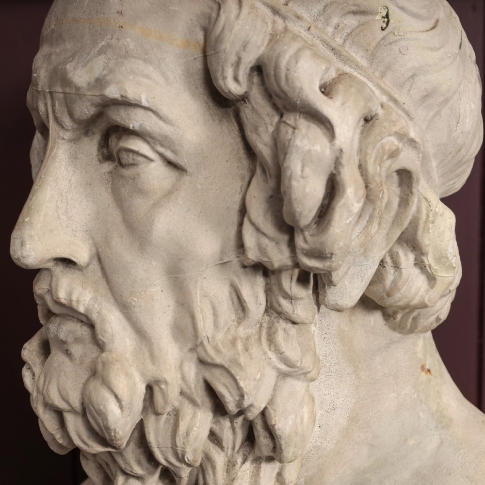 An off-white plaster bust depicting the classical Greek poet Homer with a detailed curly beard and headband, set on a flat square base. View IX