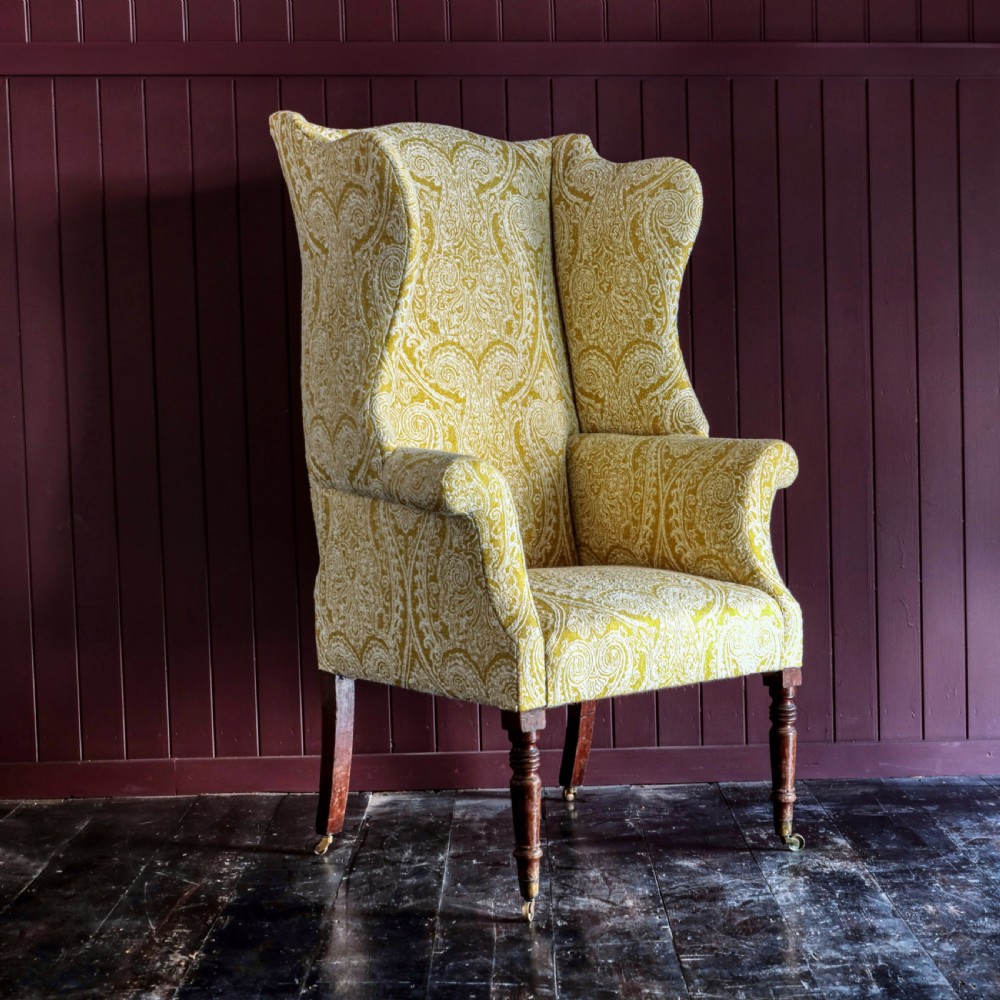 A high-backed Regency wingback armchair upholstered in patterned yellow fabric with turned mahogany front legs on brass castors.
