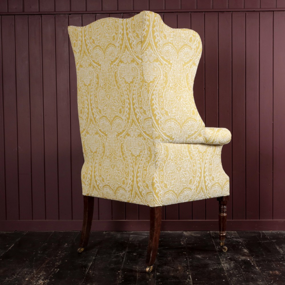 A high-backed Regency wingback armchair upholstered in patterned yellow fabric with turned mahogany front legs on brass castors. View IX