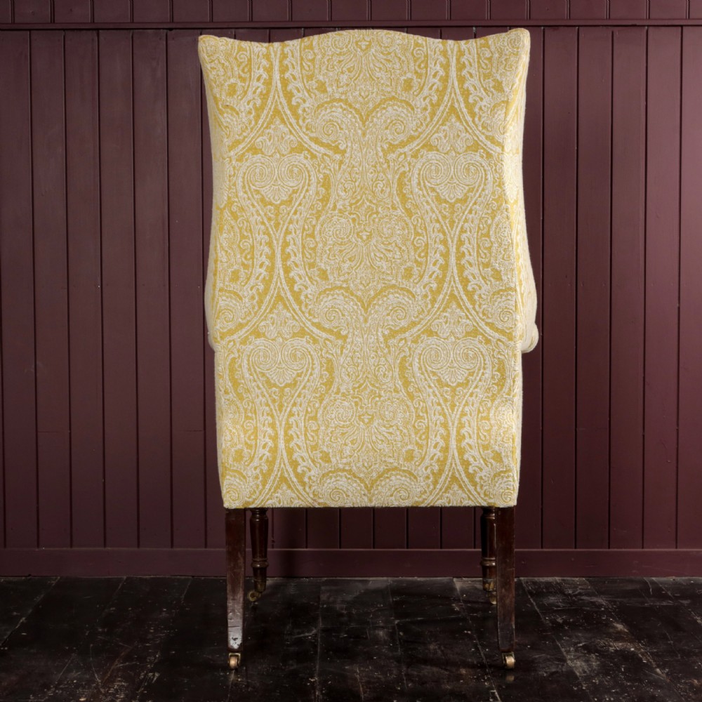 A high-backed Regency wingback armchair upholstered in patterned yellow fabric with turned mahogany front legs on brass castors. View X