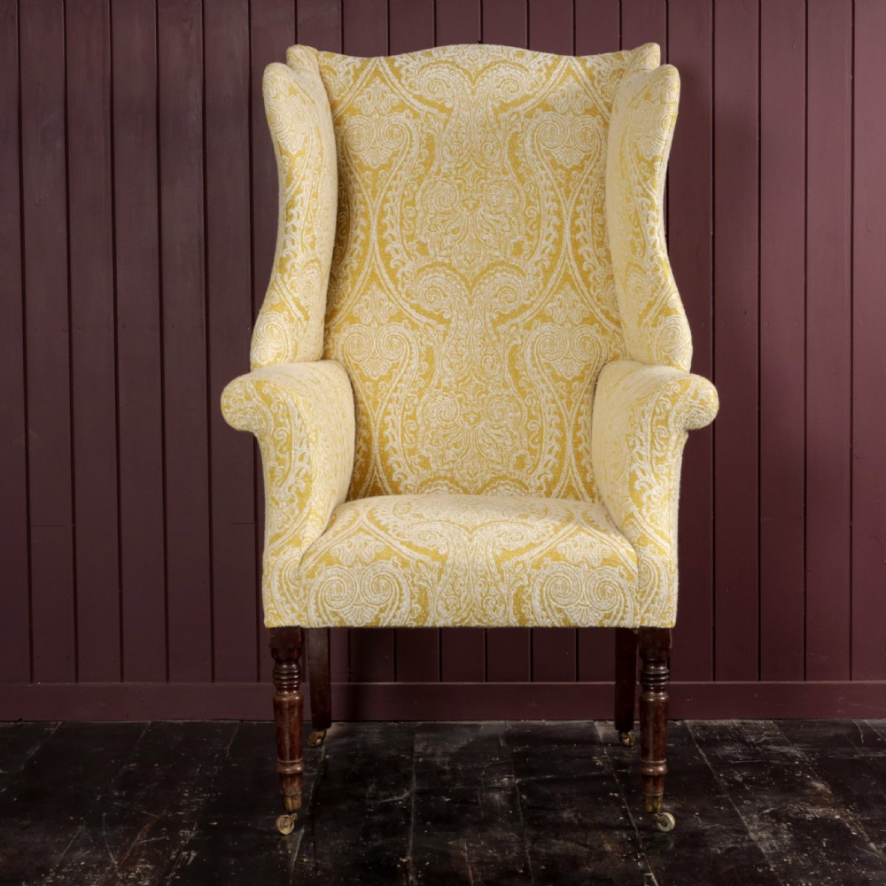 A high-backed Regency wingback armchair upholstered in patterned yellow fabric with turned mahogany front legs on brass castors. View XII