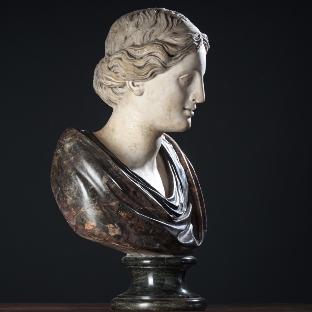 A carved marble profile bust depicting a woman with wavy hair gathered in a bun, wearing dark draped robes, set upon a turned marble socle.