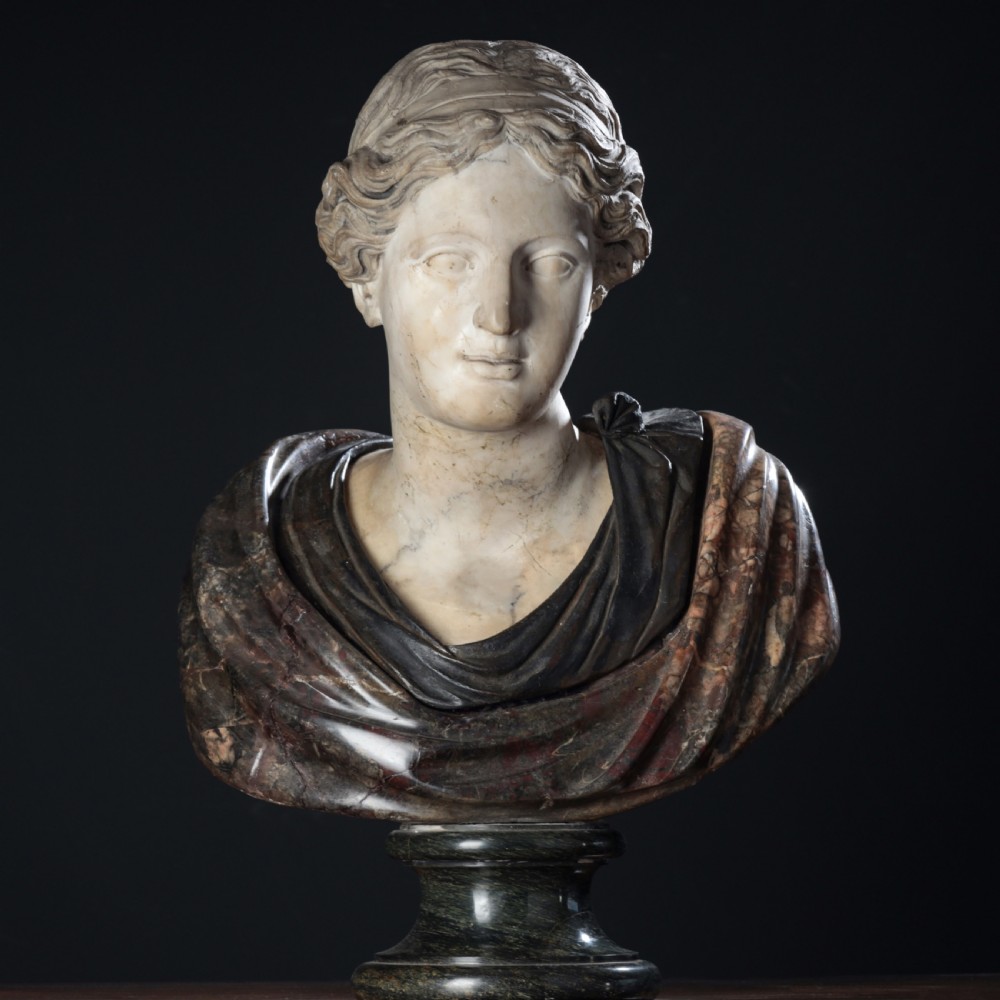 A carved marble profile bust depicting a woman with wavy hair gathered in a bun, wearing dark draped robes, set upon a turned marble socle. View II