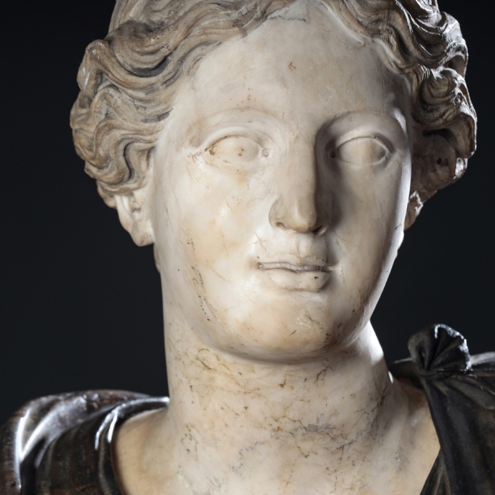 A carved marble profile bust depicting a woman with wavy hair gathered in a bun, wearing dark draped robes, set upon a turned marble socle. View III