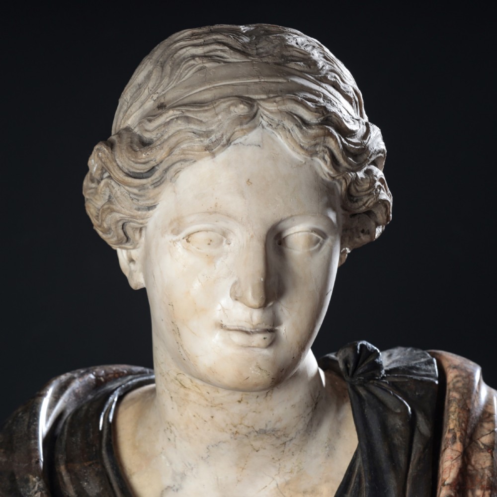 A carved marble profile bust depicting a woman with wavy hair gathered in a bun, wearing dark draped robes, set upon a turned marble socle. View IV