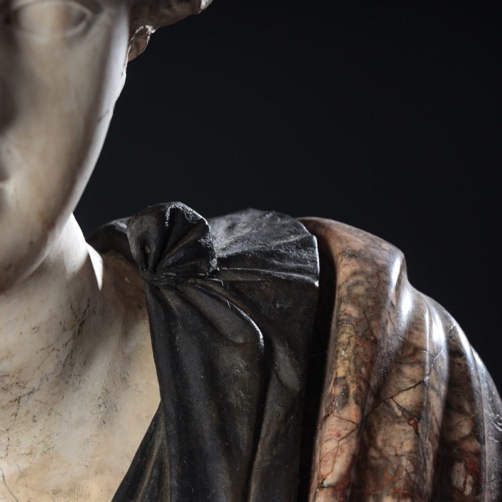 A carved marble profile bust depicting a woman with wavy hair gathered in a bun, wearing dark draped robes, set upon a turned marble socle. View V