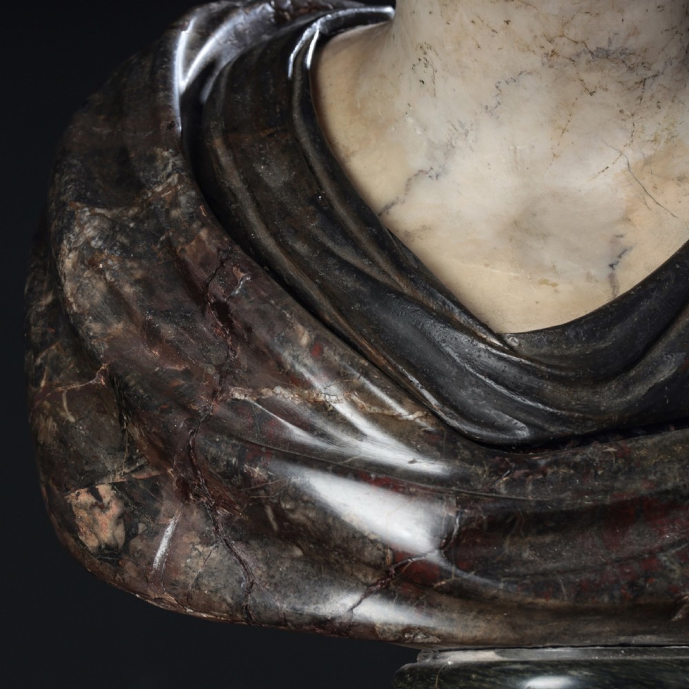A carved marble profile bust depicting a woman with wavy hair gathered in a bun, wearing dark draped robes, set upon a turned marble socle. View VI