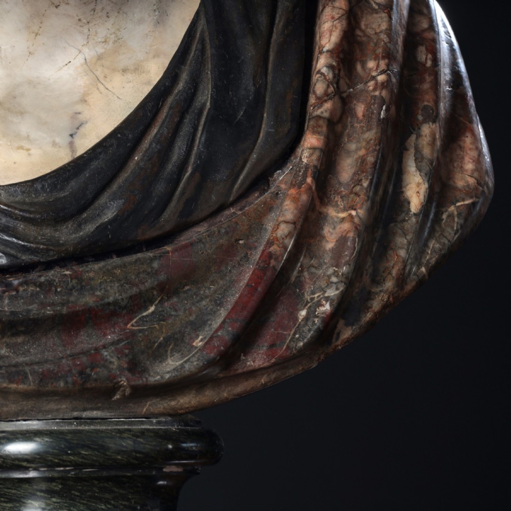 A carved marble profile bust depicting a woman with wavy hair gathered in a bun, wearing dark draped robes, set upon a turned marble socle. View VII