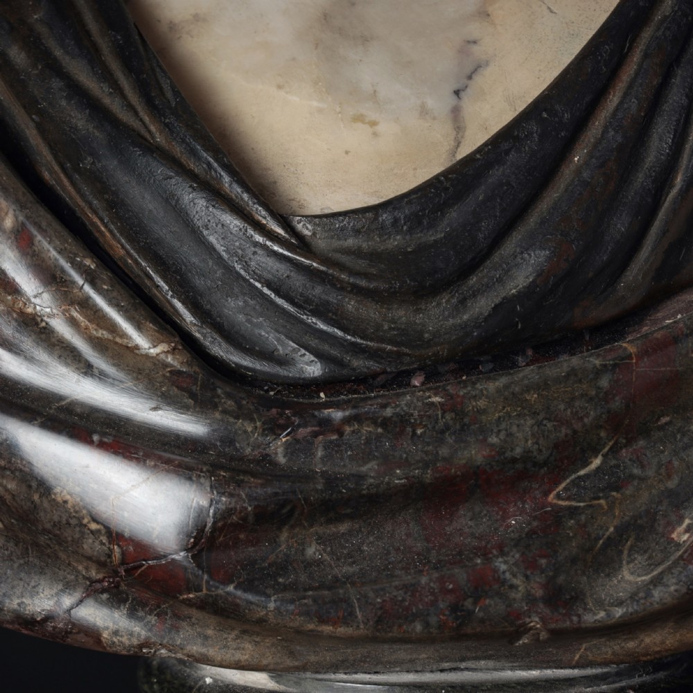 A carved marble profile bust depicting a woman with wavy hair gathered in a bun, wearing dark draped robes, set upon a turned marble socle. View VIII